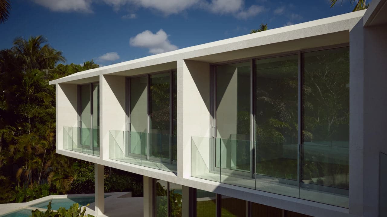 Angular white volume and glass roofline of a modern Miami residence against sky and mature tree canopy, no people in frame