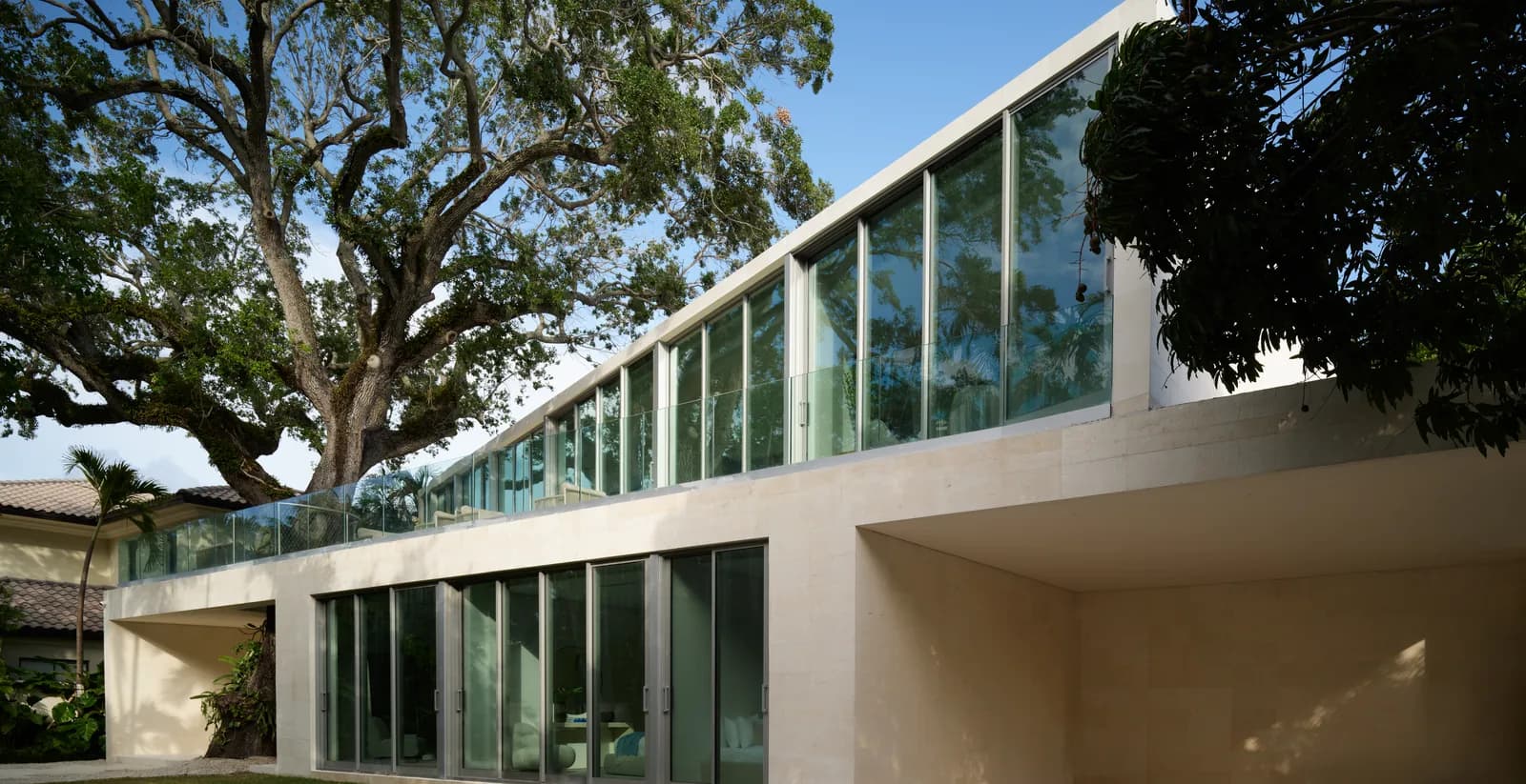 Exterior of the Onaway Residence in Coconut Grove, Miami: a white stucco pavilion wing with floor to ceiling glass, set beneath a mature oak canopy