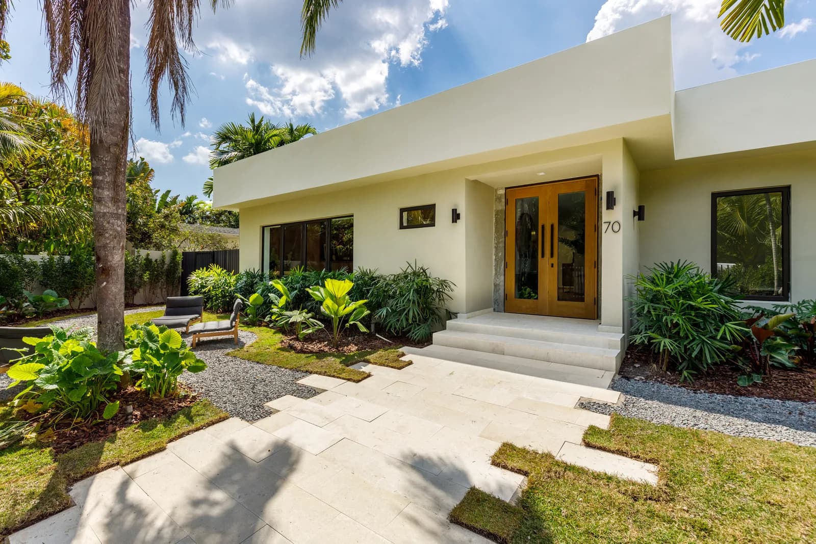 White stucco Luxom residence entry with a double oak door, black-framed glass windows and a canopy roof shading a stone walkway lined with tropical plants