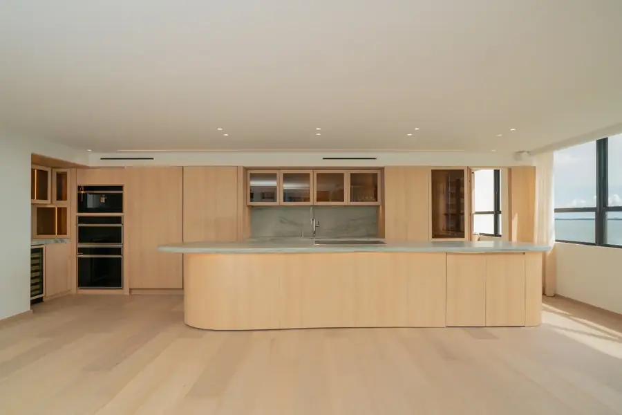 A curved kitchen island in white oak cabinetry with a honed stone waterfall countertop at the Grove Isle Residence in Miami, with floor to ceiling windows framing the water beyond